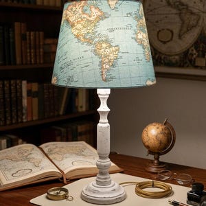 May include: A table lamp with a white, distressed-finish wooden base and a tapered lampshade featuring a vintage-style world map print in shades of blue, tan, and cream. The lamp emits a soft light from beneath the map-patterned fabric shade.