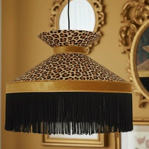 Può includere: Un paralume con stampa leopardata, finiture in velluto dorato e frange nere. Il paralume ha una forma svasata ed è sospeso a un cavo nero. Lo sfondo comprende specchi dorati ornati.