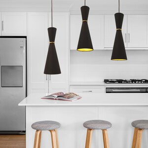 May include: Three black pendant lights with gold accents hang over a white kitchen island with three gray bar stools. A cookbook is on the counter.
