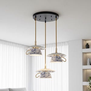 Może przedstawiać: Trzy lampy wiszące o unikalnym wzorze filiżanki do herbaty. Każda lampa ma biało-niebieski klosz z kwiatowym wzorem i złotym uchwytem. Lampy wiszą z czarnej płyty sufitowej ze złotymi przewodami.