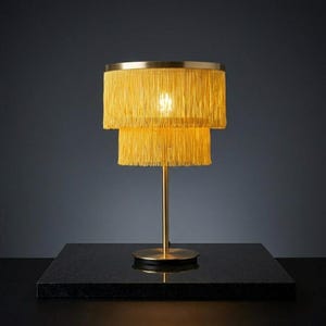 Op de afbeelding: Een tafellamp met een tweekleurige gele franje lampenkap en een frame met een metallic gouden afwerking. De lamp heeft een slanke goudkleurige centrale stang en een ronde gouden voet, wat zorgt voor een gestructureerd decoratief armatuur.