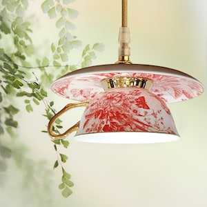 Op de afbeelding: Een unieke hanglamp gemaakt van een kop en schotel. De kop heeft een rood bloemen- en vlinderpatroon op een witte achtergrond, met een gouden oor en rand. De schotel is ook versierd met rode bloemmotieven. De lamp hangt aan een gouden snoer.