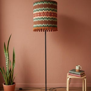Może przedstawiać: Lampa podłogowa z wzorzystym abażurem. Abażur ma powtarzający się zygzakowaty wzór w kolorze zielonym, czerwonym i białym. Lampa ma czarny metalowy stojak i podstawę. Abażur jest wykończony pomarańczowymi frędzlami. Lampa ma około 150 cm wysokości.