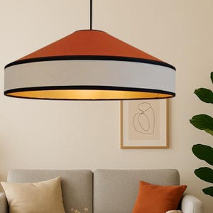 Może przedstawiać: Nowoczesna lampa wisząca z górą w kolorze spalonej pomarańczy, białym pasem i czarnym obramowaniem. Wnętrze klosza jest złote. Światło wisi nad szarą sofą z pomarańczowymi i beżowymi poduszkami.