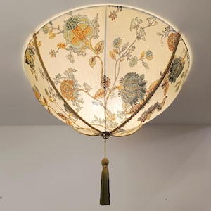 Puede incluir: Una lámpara de techo decorativa con un estampado floral. La lámpara presenta una tela color crema con un patrón repetido de flores en naranja, verde y azul. Está adornada con una cuerda dorada y tiene una borla colgando de la base.