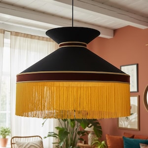Op de afbeelding: Zwarte hanglamp met een brede, conische kap. De kap heeft een bordeauxrode band met gouden rand en een lange, gele franje. Het licht hangt aan een zwart snoer.