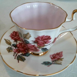 May include: A white teacup and saucer with a pink interior and gold trim. The saucer features a floral design with pink roses and green leaves.