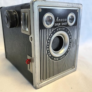 May include: Vintage Ansco Shur Shot camera. The black camera has a silver-colored faceplate with the text "Ansco Shur Shot" and "Made in U.S.A." The camera has a lens, viewfinder, and a red button. The camera is a box-shaped camera.