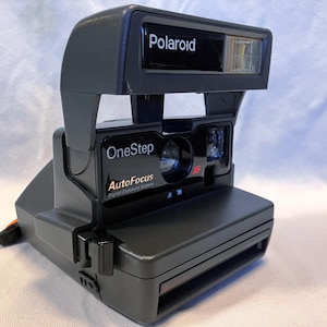 May include: A dark gray Polaroid OneStep AutoFocus instant camera featuring a prominent flash unit on top, a central lens, and a rectangular film ejection slot at the base. The front panel displays the text Polaroid, OneStep, and AutoFocus.
