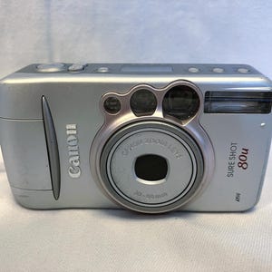 May include: Silver Canon Sure Shot 80u digital camera with a zoom lens. The camera has a built-in flash and a wrist strap. The lens has a 28-90mm range.
