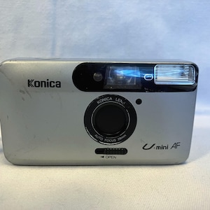 May include: Silver Konica U-mini AF 35mm film camera. The camera has a black lens and flash, with the Konica logo on the front. The lens reads "Konica Lens Auto Focus 28mm". The camera has a black and gray strap.