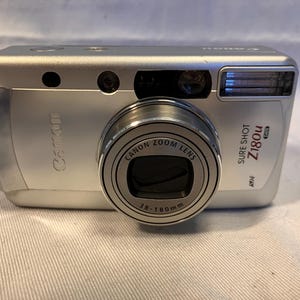 May include: A silver Canon Sure Shot Z180u point-and-shoot film camera with a 38-180mm zoom lens. The front of the camera features the Canon logo, a built-in flash, a viewfinder, and a wrist strap attached to the left side of the camera body.