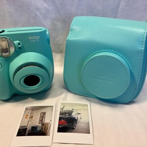May include: A turquoise Instax Mini 7S instant camera with a matching turquoise protective carrying case and two developed instant film photos. One photo shows an interior room with a lamp, and the other shows a dark car parked in a garage.
