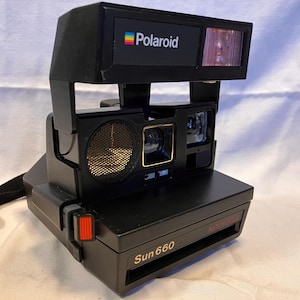 May include: A black Polaroid Sun 660 instant camera featuring a rectangular body, a built-in flash unit, a square lens, and a circular sonar autofocus sensor. The camera includes a black wrist strap and displays the Polaroid logo and Sun 660 text.