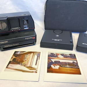 May include: A black Polaroid Impulse SE camera with a red accent, a black case, a Polaroid receiver, and a Polaroid accessory. Two Polaroid photos are also included, showing interior and exterior scenes.