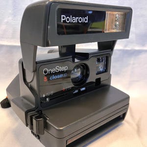 May include: A dark gray Polaroid OneStep Closeup instant camera featuring a built-in flash unit at the top, a central lens, and a rectangular body. The front panel displays the text Polaroid, OneStep, and Closeup in white and blue lettering.