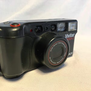 Peut inclure: Appareil photo Vivitar 3001z noir avec objectif zoom. L'appareil photo a un bouton rouge et un flash. L'objectif porte l'inscription "Vivitar Series 1 Auto Focus Lens" et "38-80mm Zoom Macro Focus Japan". Une sangle marron est attachée.