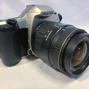 May include: A silver and black Pentax F-30 camera with a large black lens. The lens has the text "ASPHerical" and "Jantaray" printed on it. The camera is on a white surface and has a black strap.