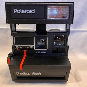 May include: A black Polaroid OneStep Flash instant camera featuring a rectangular flash unit, a central lens, and a red vertical stripe accent on the front. The camera body displays the text Polaroid and OneStep Flash with distance settings.