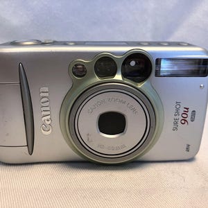 May include: A silver Canon Sure Shot 90u Date point-and-shoot film camera with a retractable zoom lens. The front features a Canon Zoom Lens 38-90mm, a built-in flash, and a light green accent ring surrounding the lens housing.