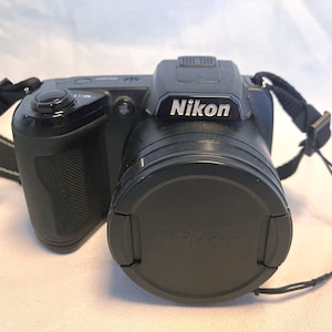 May include: A black Nikon digital camera featuring a textured grip, a prominent lens with a protective cap, and a black neck strap attached to the sides. The camera body displays the Nikon brand name in white lettering on the top housing.