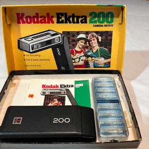 May include: A vintage Kodak Ektra 200 camera outfit in its original box. The box is yellow with black and red text. The camera is black with silver accents. The kit includes the camera, a case, film, and paperwork. The box features an image of the camera and two people.