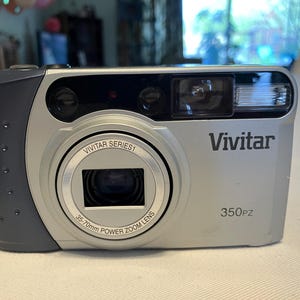 May include: Silver and gray Vivitar 350PZ digital camera with a 35-70mm power zoom lens. The camera has a built-in flash and a black strap. The Vivitar Series 1 logo is visible on the lens.