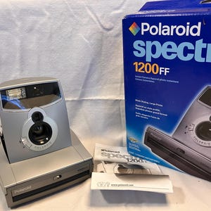 May include: A silver Polaroid Spectra 1200FF instant camera with a matching blue product box and a white instruction manual. The camera features a prominent lens, a flash unit, and the Polaroid logo on the front of the device body.
