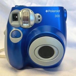 May include: A blue Polaroid 300 instant camera with a silver lens and flash. The camera has a light gray accent on the side and a matching strap. The camera has a 60mm focus range.