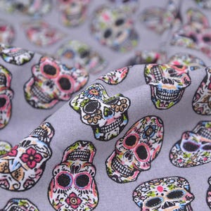 May include: Grey fabric patterned with vibrant sugar skulls. Each skull is adorned with floral and decorative motifs in pink, green, blue, and yellow. Ideal for crafting, the fabric has a detailed and colourful design.