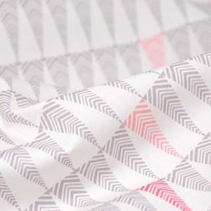 May include: Close-up of white fabric with a repeating geometric pattern of gray and pink arrow shapes. The fabric is draped, creating soft folds and shadows. The design is modern and minimalist.