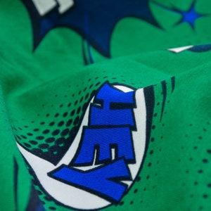Könnte beinhalten: Grüner Stoff mit einem weißen und blauen Comic-Druck. Der Druck zeigt das Wort "HEY" in großen blauen Buchstaben.