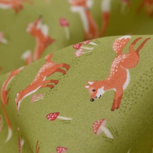 May include: Close-up of green fabric with a repeating pattern of orange foxes and small red and white mushrooms. The foxes are in various poses, with white accents and black eyes. The fabric has a textured appearance.
