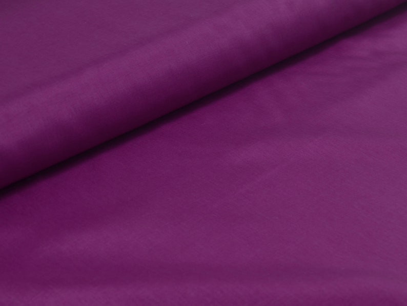 May include: A smooth, solid purple fabric with a slight sheen. The fabric is folded in half, with one side lying flat and the other side folded over it.