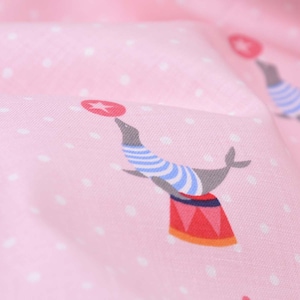 May include: Pink fabric with a white polka dot pattern featuring a gray seal balancing a red star on its nose. The seal is wearing a blue and white striped shirt and is standing on a red and white striped drum.