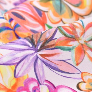 Op de afbeelding: Close-up van een bloemenstof met een levendig patroon. Het ontwerp toont grote, gestileerde bloemen in tinten paars, oranje, roze en geel tegen een witte achtergrond. De stof is gevouwen en toont de textuur en details van de print.