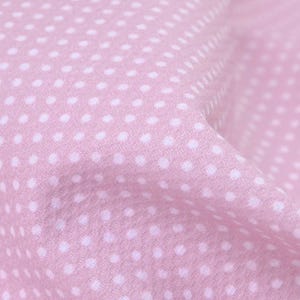 May include: Close-up of a pink fabric with a repeating pattern of small white polka dots. The fabric is soft and slightly wrinkled, with the polka dots evenly spaced across the surface. The overall effect is delicate and feminine.