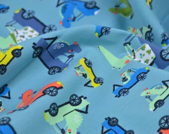 Puede incluir: Tela azul con un patrón repetido de coches, camiones y dinosaurios coloridos. Los coches son amarillos, rojos y azules. Los camiones son azules y verdes. Los dinosaurios son verdes y azules.