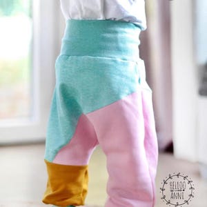 May include: Colorful toddler pants in shades of pink, teal, and mustard yellow. The pants have a high waistband and are made of soft fabric. The pants are designed with a unique color-block pattern.