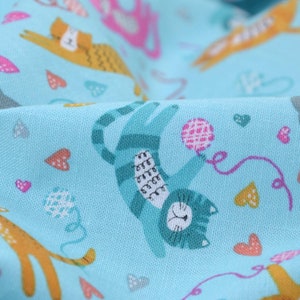 Peut inclure: Un tissu bleu avec un motif répétitif de chats jouant avec des pelotes de laine et des cœurs. Les chats sont de différentes couleurs, notamment turquoise, orange et gris.