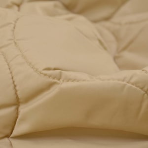 May include: Close-up of a beige quilted fabric. The fabric has a diamond pattern created by stitched seams. The material appears soft and slightly textured, with subtle variations in colour and shadow.