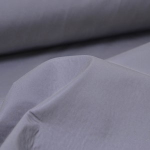 Op de afbeelding: Close-up van een gladde, grijze stof met een subtiele glans. De stof is gevouwen op een manier die een zachte, afgeronde vorm creëert.