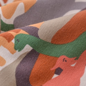Op de afbeelding: Close-up van een stof met een kleurrijk abstract patroon met dinosaurussen in tinten oranje, groen, bruin en wit.