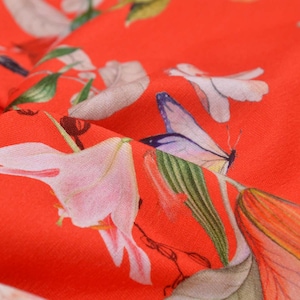 Puede incluir: Primer plano de tela roja vibrante con estampado floral y de mariposas. El diseño presenta flores blancas y rosas, junto con mariposas en tonos morados, azules y amarillos. La tela está doblada, mostrando la textura y el patrón.