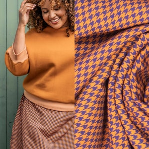 Peut inclure: Une femme portant un pull orange brûlé et une jupe à motif pied-de-poule noir et orange. La jupe est trapèze et arrive aux genoux. Le tissu est texturé et a un léger brillant. La femme sourit et a les cheveux bruns bouclés. L'arrière-plan est un mur vert avec un tissu à motif pied-de-poule drapé dessus. Le tissu est orange et violet. Le texte #betsy est visible dans le coin inférieur droit de l'image.