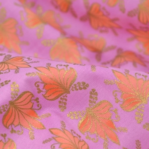 Könnte beinhalten: Lilafarbener Stoff mit einem sich wiederholenden Muster aus orangefarbenen Blumenmotiven. Der Stoff hat einen dezenten Glanz und eine weiche Textur.