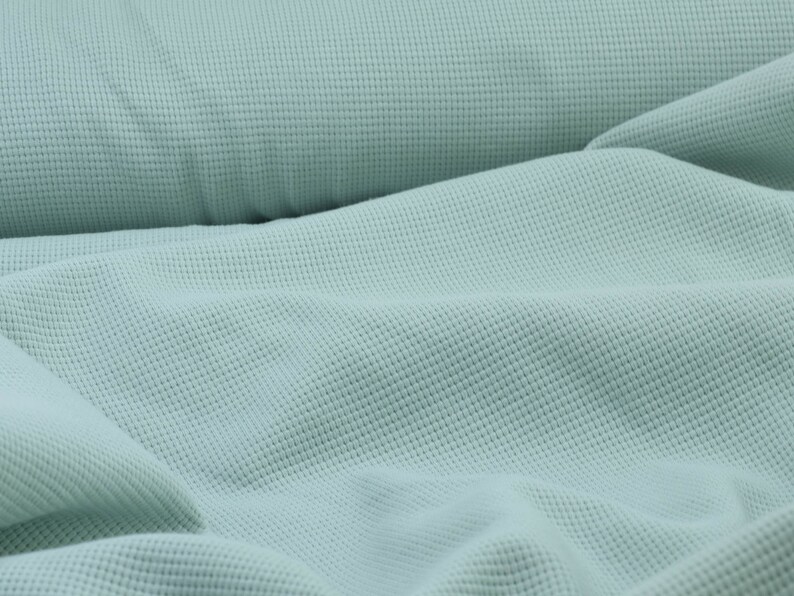 May include: A close-up of a light blue waffle knit fabric. The fabric is soft and textured, with a subtle sheen.
