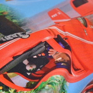 Può includere: Un'auto giocattolo rossa con strisce bianche e il testo "PAT 3" sul lato. L'auto è parzialmente coperta da una pianta verde frondosa.