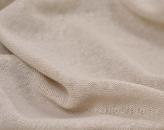 May include: Close-up of a soft, beige fabric with a textured knit pattern. The fabric is folded and draped, creating a smooth, flowing surface.