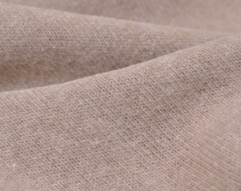 Pode incluir: Close-up de um tecido de malha bege com textura visível e variações sutis de cor. O tecido está dobrado, criando sombras suaves e destacando a textura do material. A imagem mostra os detalhes do têxtil.